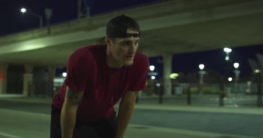 A tired athletic man breathes heavily as he stands with his hands on his knees on a road at night