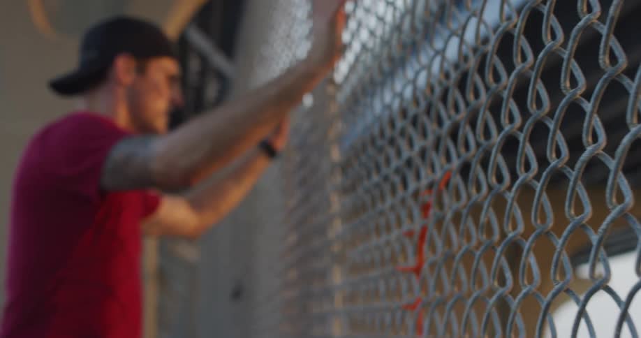 An athletic man looks sad as he hold onto and leans on a metal fence