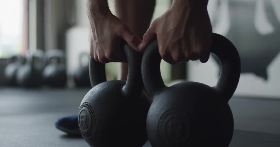 A pair of hands picks up two kettlebells from the floor