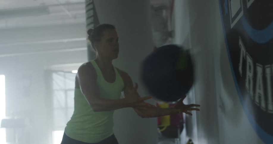 A woman exercises with a medicine ball by bouncing it on the wall in a gym