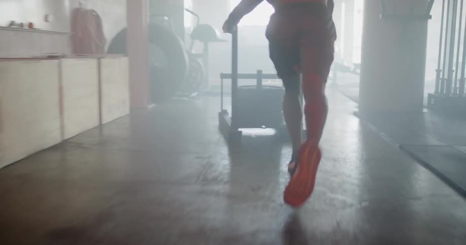 A man in a gym running while pushing a cart loaded with weights then turning it around