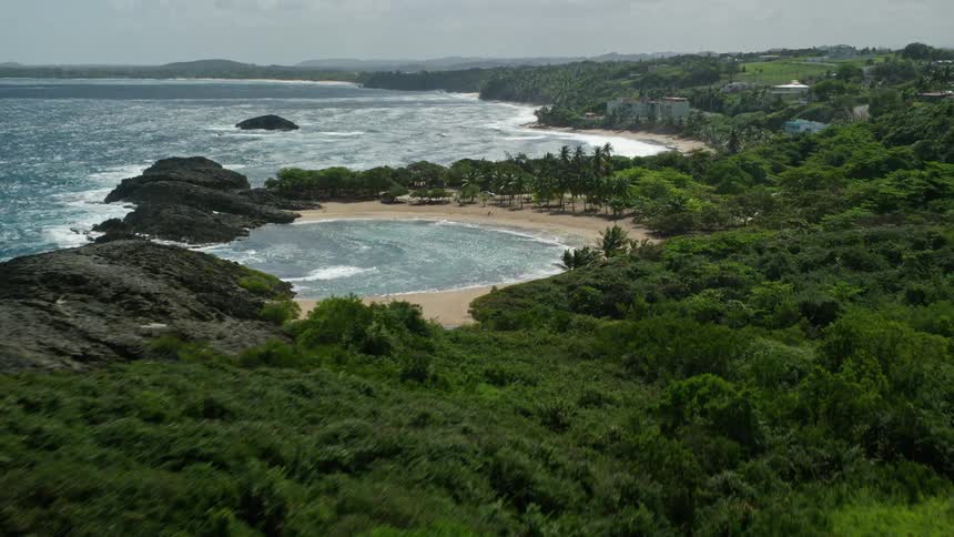 A coastal region with rocks and vegetation and further on to a small bay with a circular beach and tropical trees