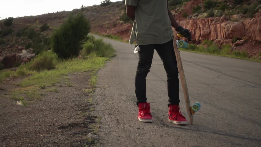 Man standing on side of road with skateboard leaning on his leg