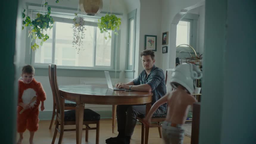 A father sits in the kitchen while his sons run around playfully