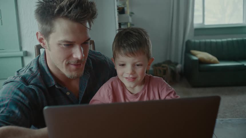 A man and a young boy sit at a table laughing at a laptop