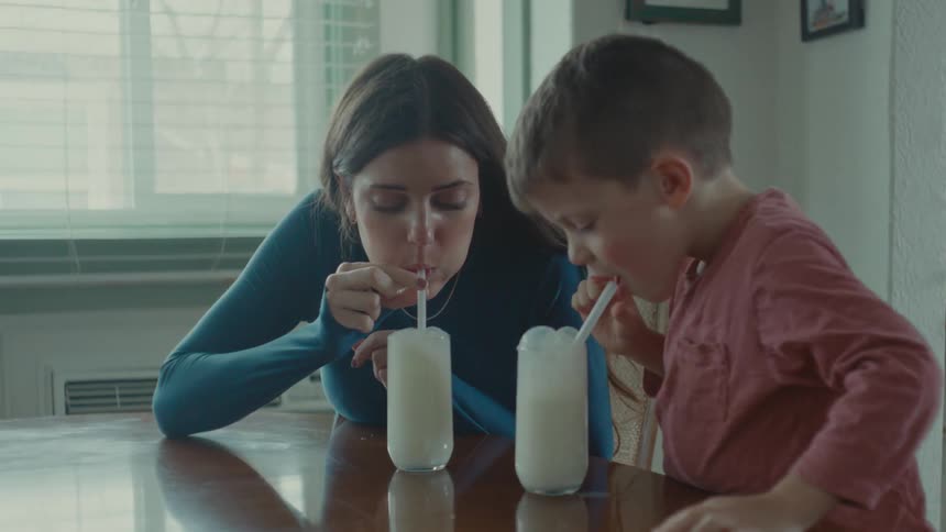 A woman and a young boy sit at a kitchen table blowing bubbles from their respective glasses of milk spilling it while laughing