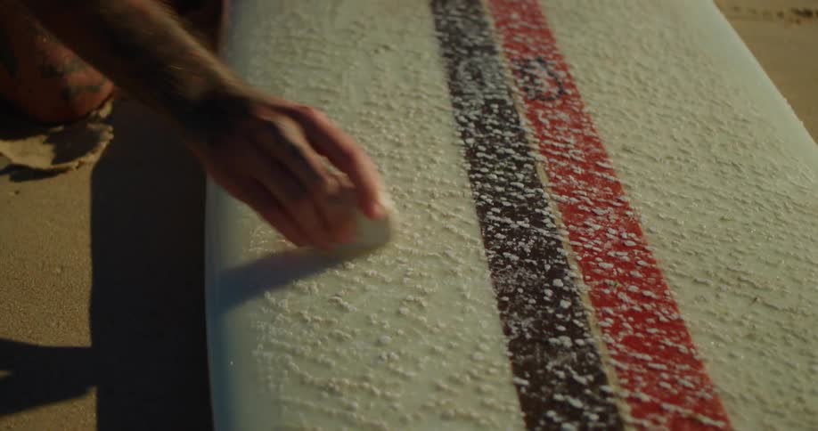 Hands of a person applying paraffin to a board that is on the beach sand