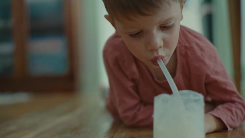 A young boy is lying on the floor blowing bubbles onto a glass of milk through a straw