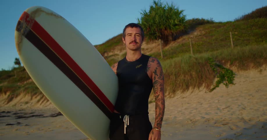 Young tattooed man holding a longboard on the beach