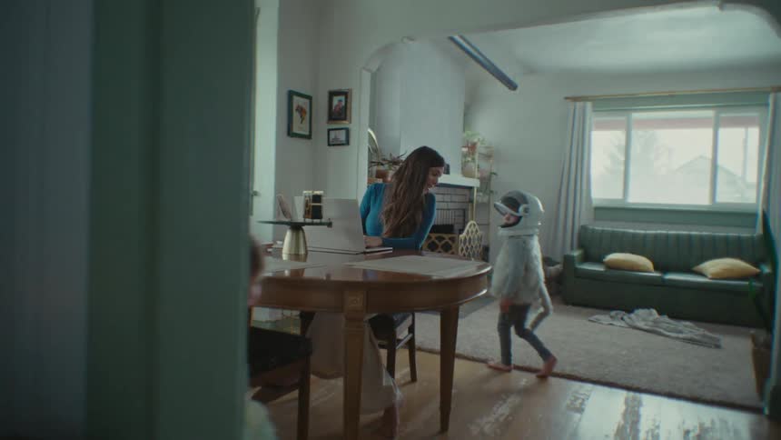 Mother sitting at a table working on her laptop and her two children walking around the table and her