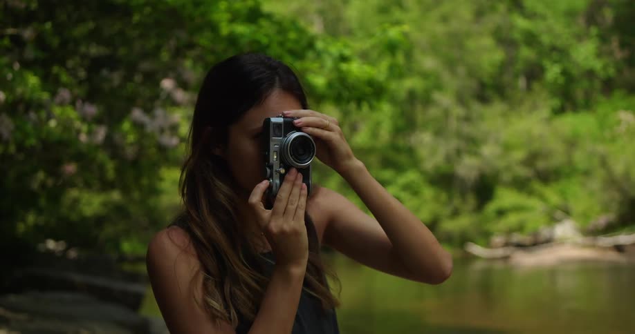 A woman near a pond in a forest takes a picture with a camera