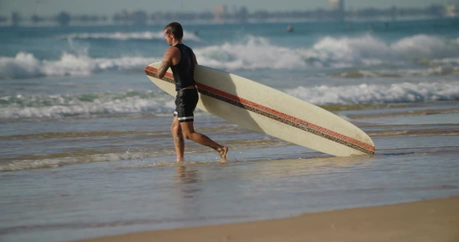 A young man runs along the beach and picks up his surfboard under clear skies