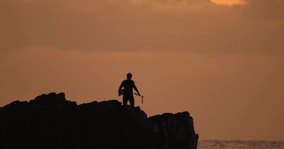 Diver carrying flippers stands on shore over water silhouetted against a red tinted sky