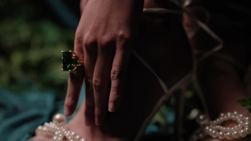 A womans hand wearing a large emerald ring passing over her strap high heel shoes decorated with pearls