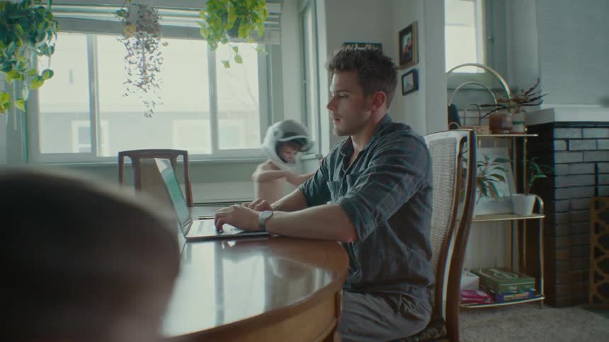 Man working on the laptop while seated indoor and his children running around the house at midday