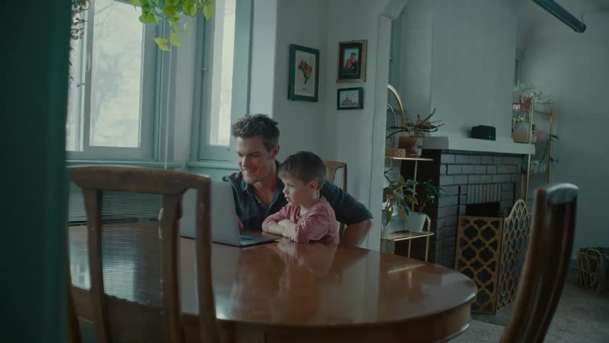 A man and a young boy sit at a table laughing at a laptop