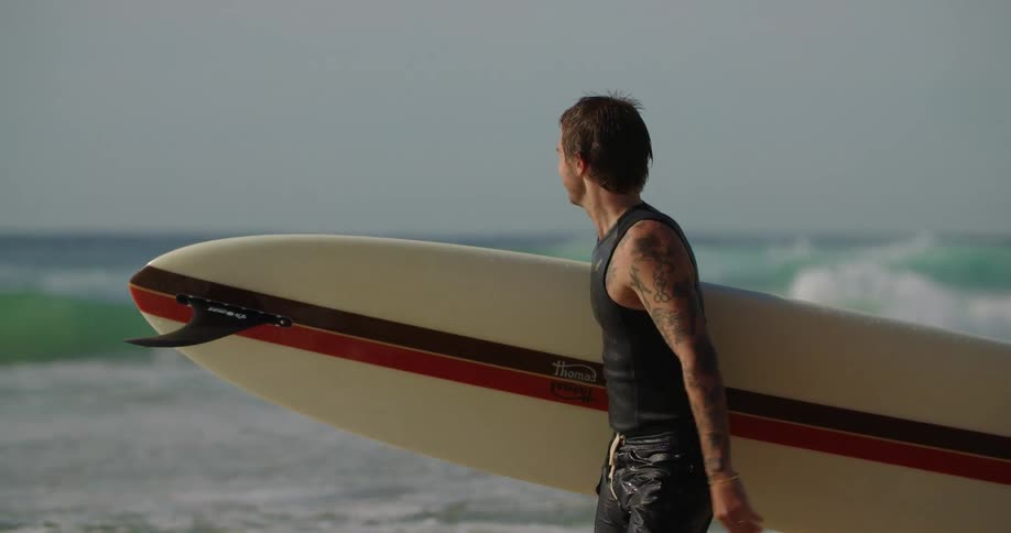 A man with a surfboard walking by the beach smiling while raising two fingers up