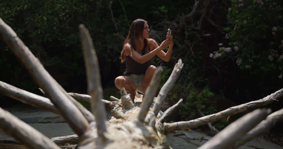 A woman squats on a log to take a picture with a smart phone