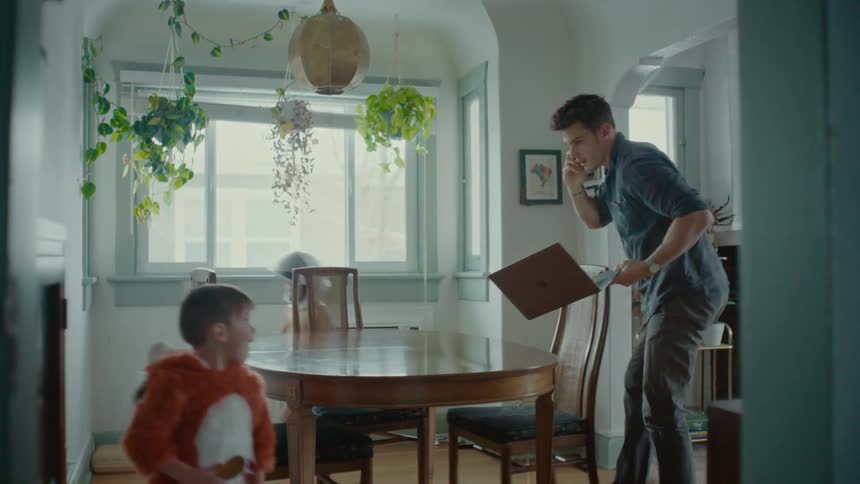 Two children run around a table where a man is sitting with a computer and phone