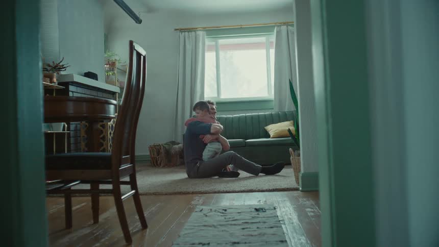 A man waits sitting on a living room floor until his young son comes running into his arms and they hug