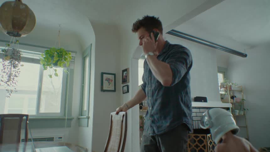 A man is working with a laptop on a dining table getting up while on a phone call but has trouble focusing as his two young sons chase each other around him