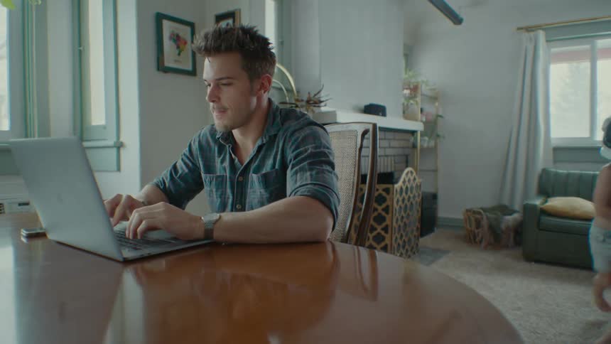 Father sitting at the table in front of a laptop and his son passing by him and the two start to play