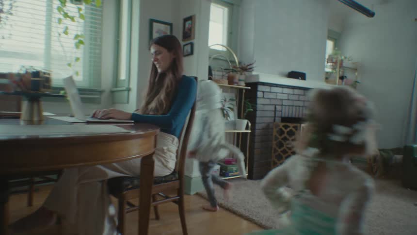 Mother working on the laptop with her two children running around her in the living room