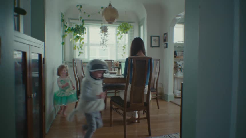 A woman tries to work at a kitchen table while her children run around her chasing each other