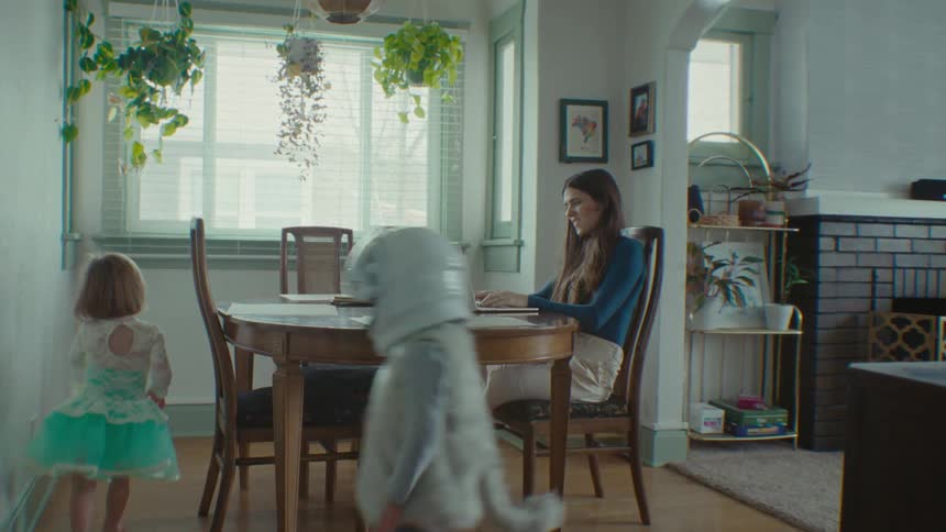 An adult woman is sitting on the chair working on the computer while two children are running around the table