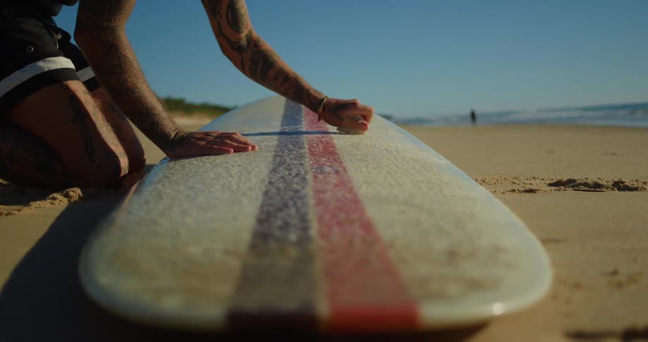 Man applying paraffin to his surfboard on the beach sand