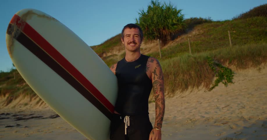 A young man stands still on the beach while holding his surfboard