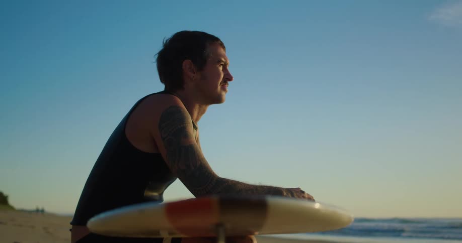 Man sitting on a beach holding a longboard looking at the sunset
