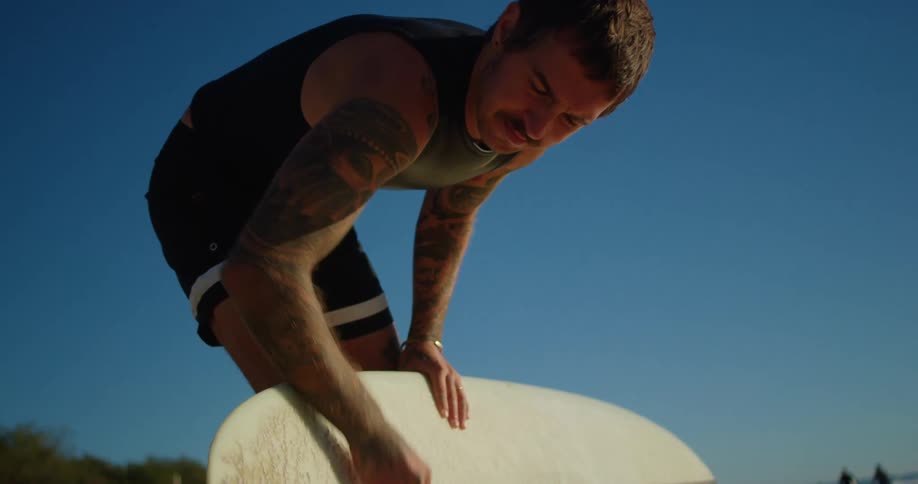 A male surfer leans over and rubs wax onto the surface of his surfboard