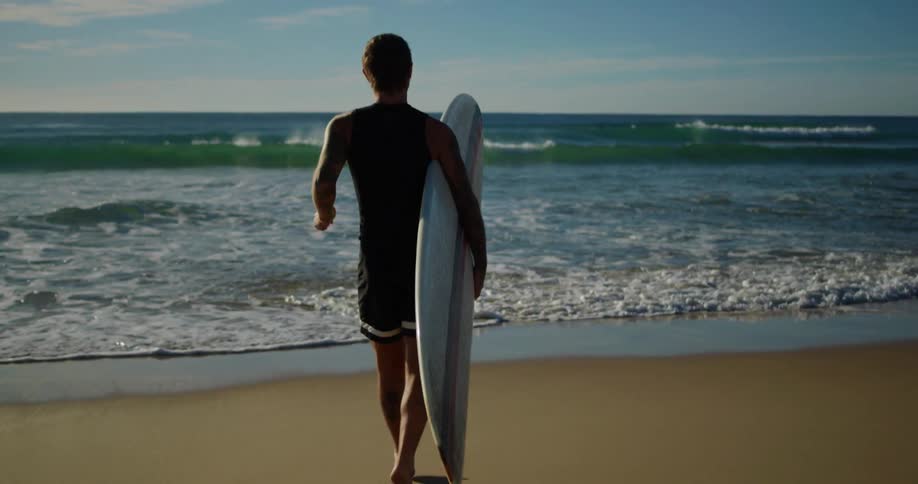 Surfer carries surfboard across beach and into water on a slightly cloudy day