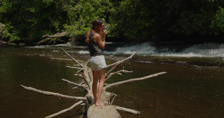 Lady take picture with a camera while standing on a fallen tree on a stream at midday