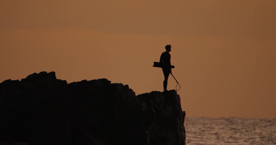 Silhouette of a person standing on rocky terrain against a sunset over the ocean