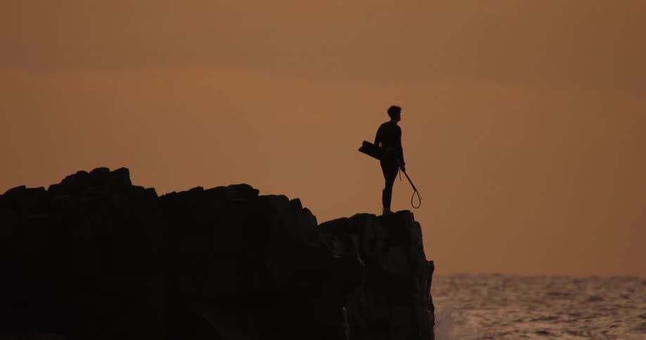 Silhouette of a person on the peak of costa with the sea wave hitting it on an orange sky day