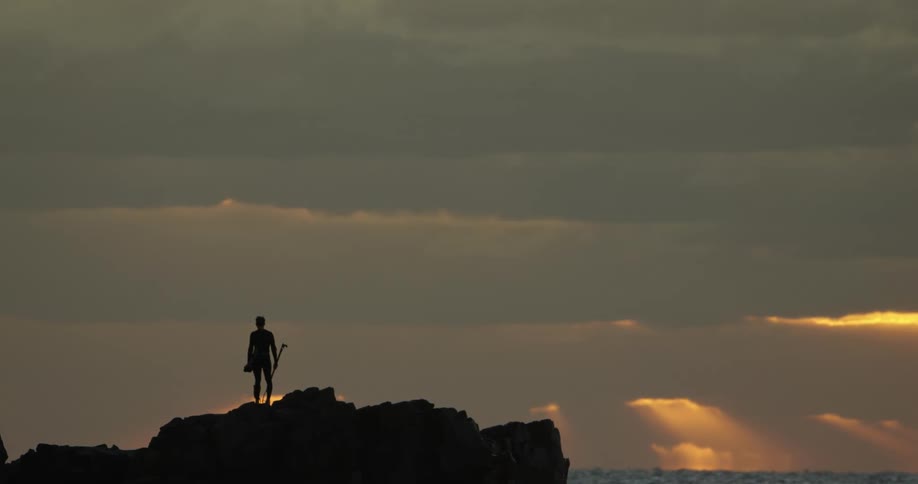 Silhouette of a male standing on a jagged coastal rock beside the sea under a sky filled with gray clouds and rays of light from a setting sun streaming down in the distance