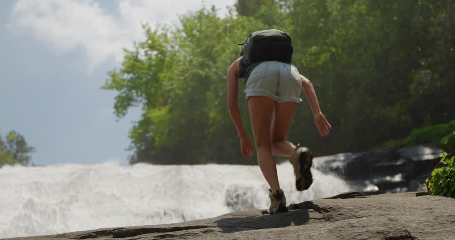 Lady climbs over a rock to watch a waterfall on a sunny day