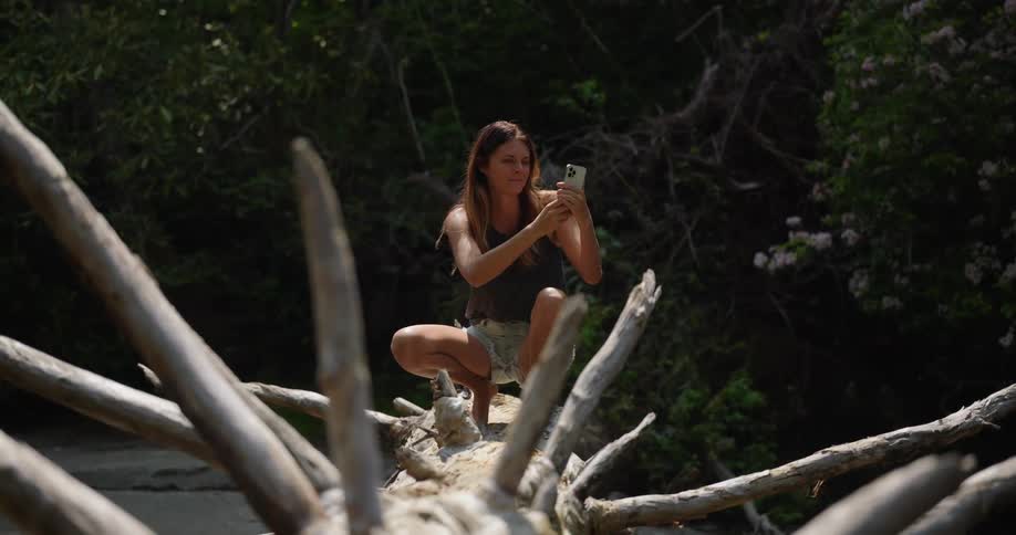 A woman couches atop a fallen tree while capturing photos on her smart phone
