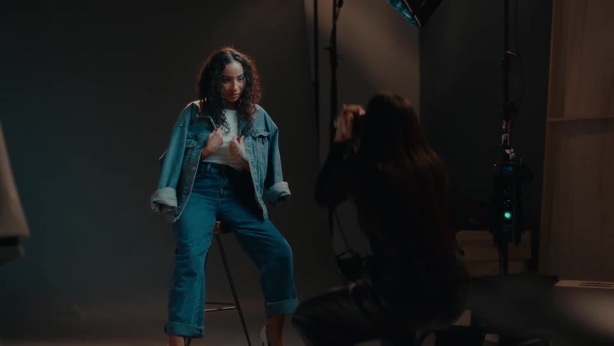 A woman is sitting wearing jeans posing for another who is taking photos of her