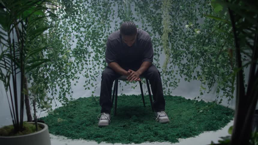 A person sitting on a stool amidst hanging vines and potted plants with hands clasped