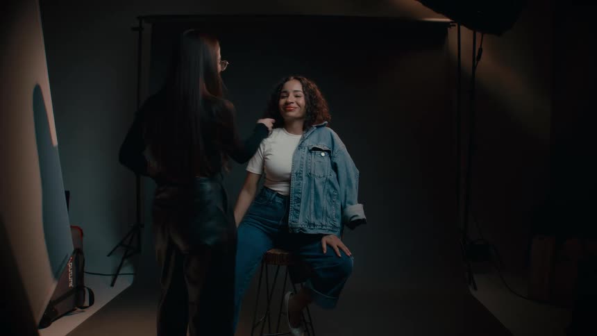 A black model on a chair smiles at a woman checking her hair