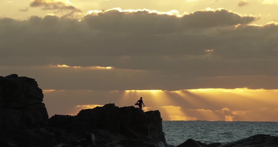 A person stands on rocky terrain by the sea against a sunset with rays of light piercing through the clouds