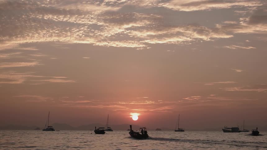 A motorboat speed out into a bay dotted with moored boats as the sun sets