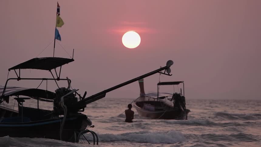 A person stands in the surf between two boats at sunset