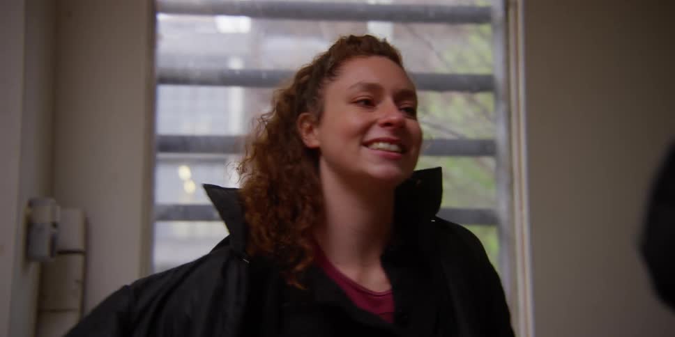 Woman smiles at friend as they put on jackets while standing near a window