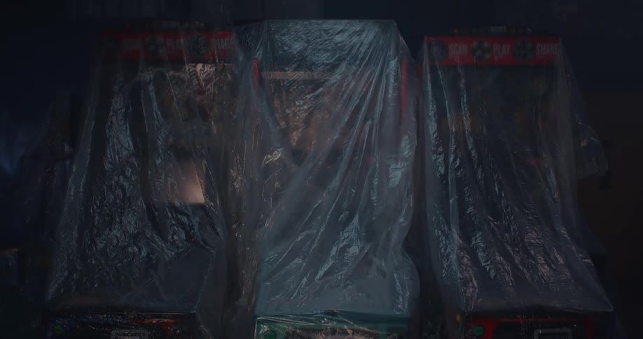 Three arcade machines in a dark room covered in a plastic bag as a beam from a flashlight passes them