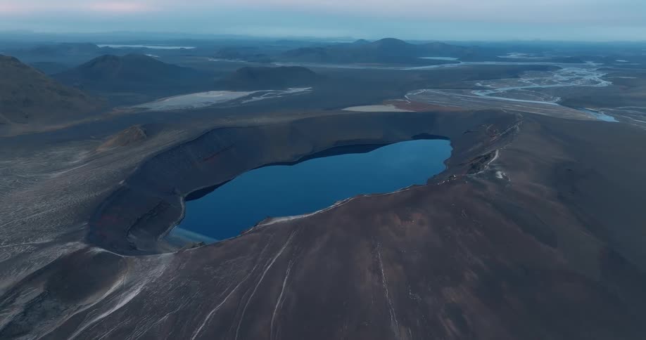 A large dark crater lake surrounded by rugged terrain with distant mountains and a cloudy sky