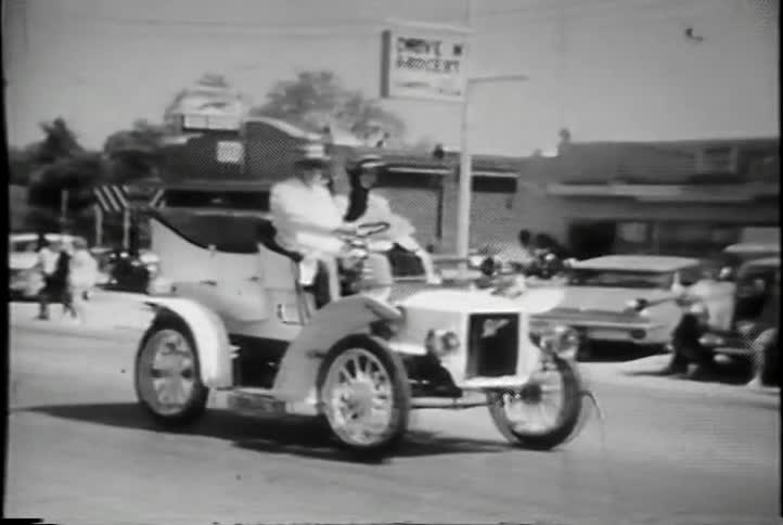 A man and woman drive a car down the street in a parade